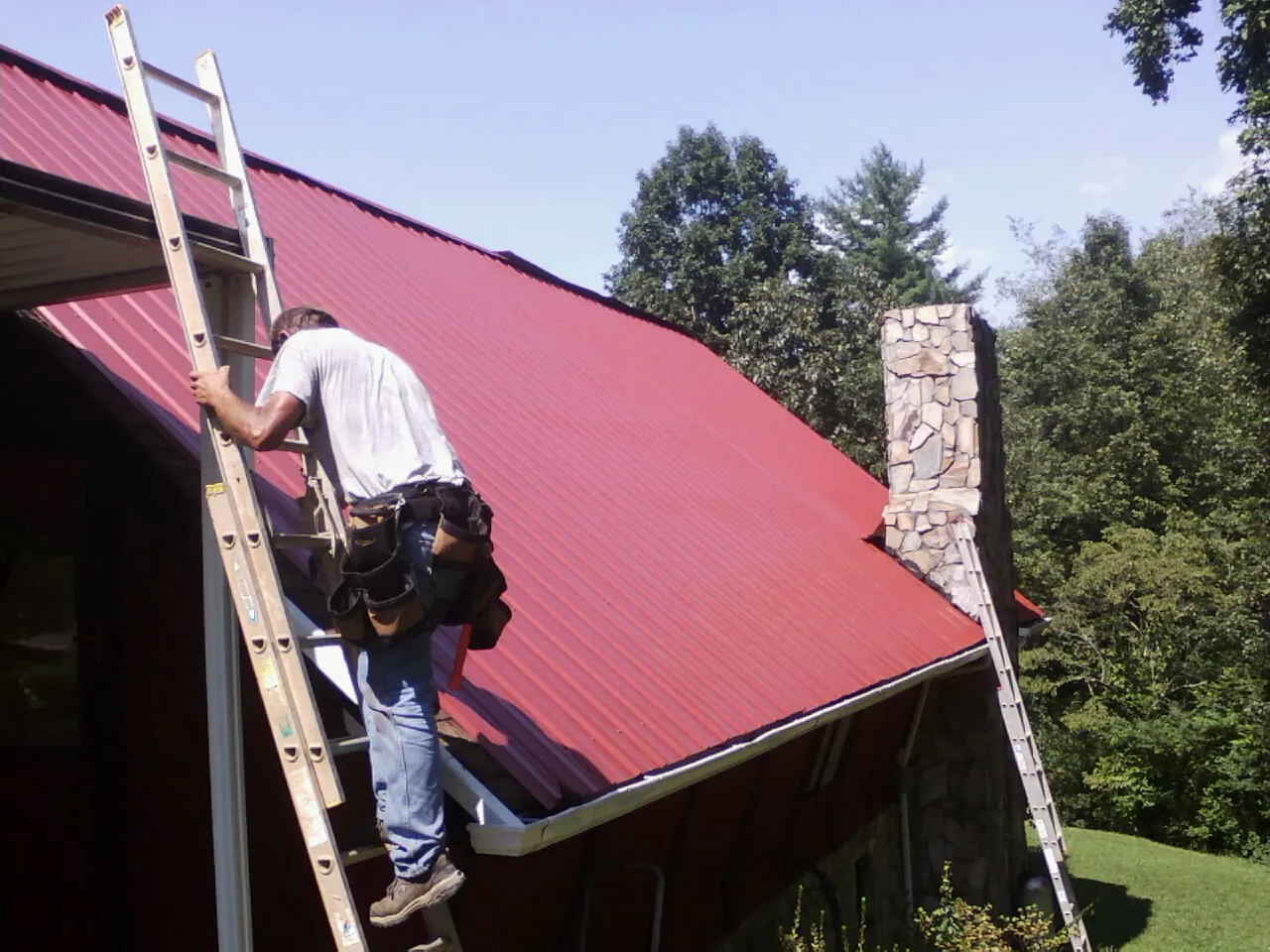 Roofer on a ladder beside a red metal roof and stone chimney — Soffit & Fascia Repair by Roofing Brothers in Liberty Hill